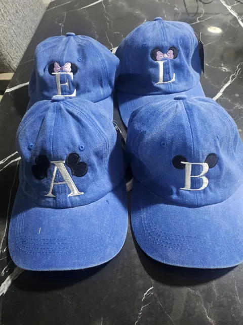 Cuatro gorras azules lavadas con una inicial bordada en blanco (E, L, A y B) y orejas de ratón bordadas, dos de ellas con moño