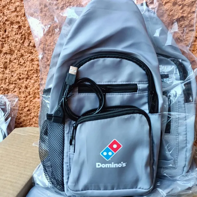 Mochila bandolera gris con el logo de Domino's bordado al frente, aún en su bolsa de empaque